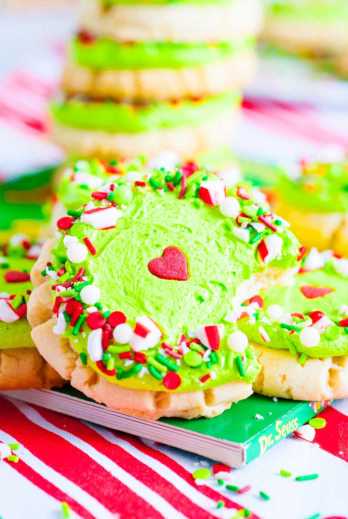 Round Grinch Crumbl Christmas Sugar Cookies with green frosting, red heart candy, and festive sprinkles sit on a Dr. Seuss book.