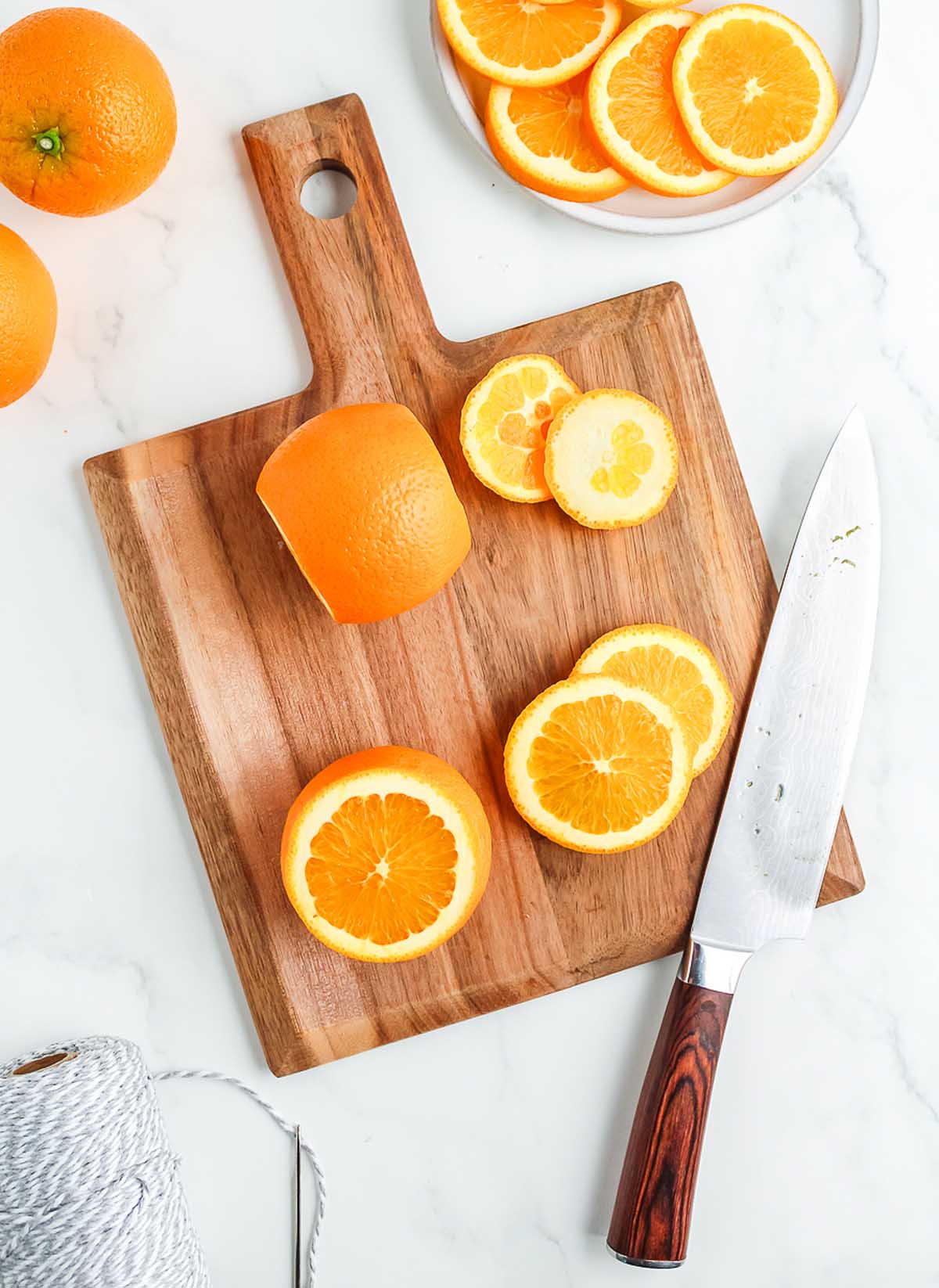 Orange slices and a whole orange on a wooden cutting board with a knife, ready to be prepared for a homemade dried orange garland, displayed on a white surface.