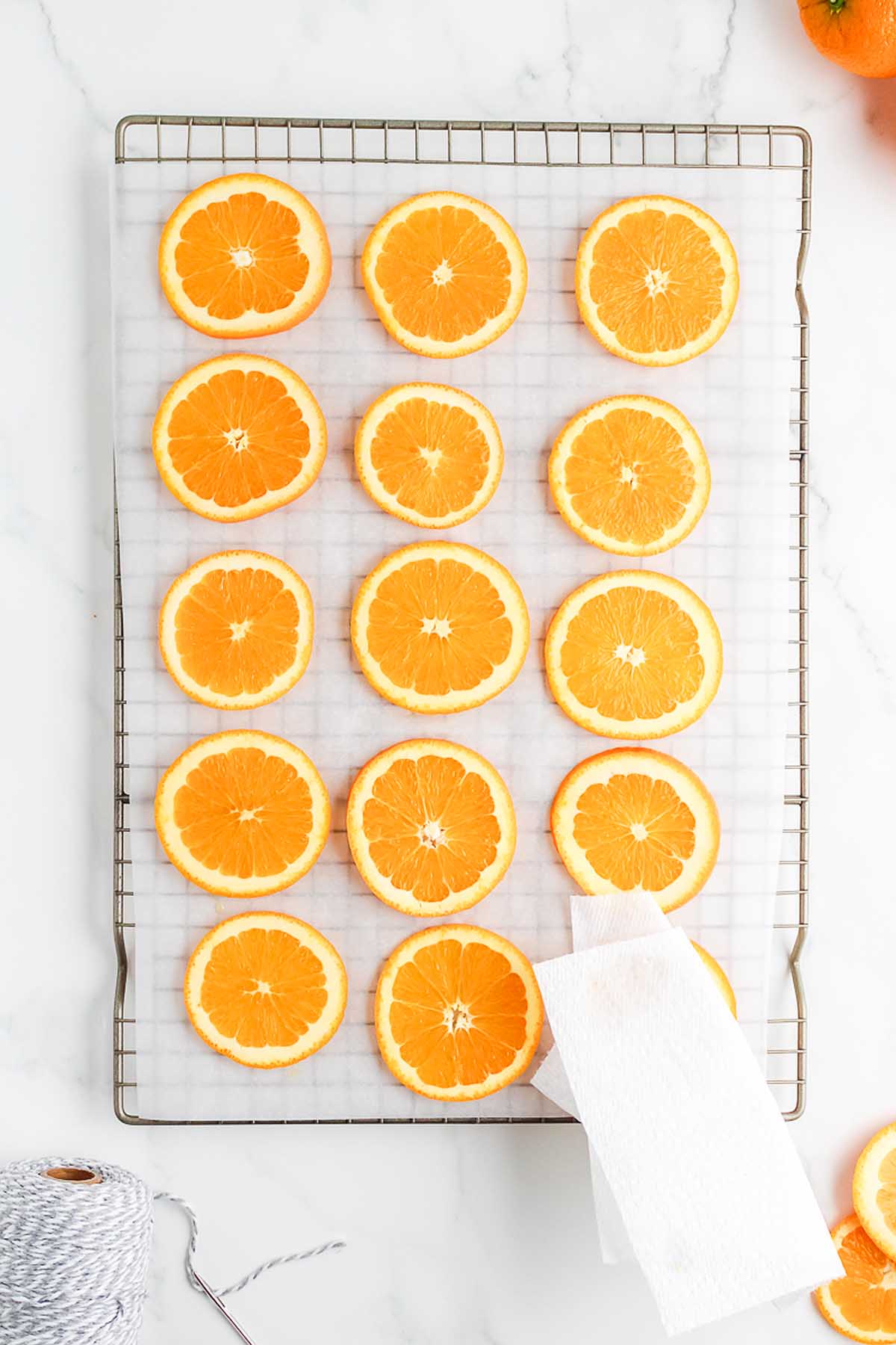 Orange slices arranged in rows on a wire rack lined with parchment paper, ready to be used for a charming Homemade Dried Orange Garland, with a paper towel at the corner.