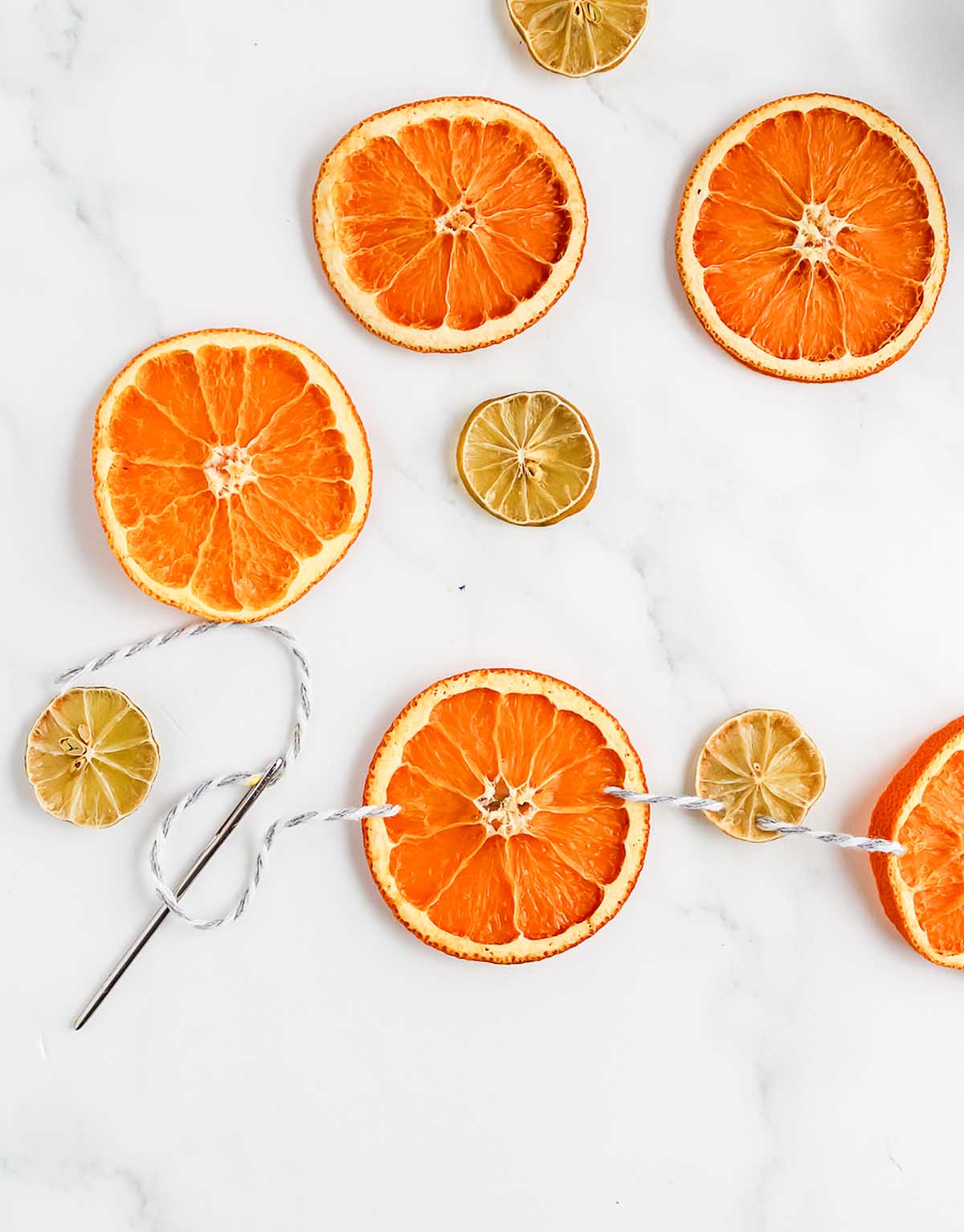 Homemade Dried Orange Garland is created by threading dried orange and lime slices onto string with a needle on a white marble surface.