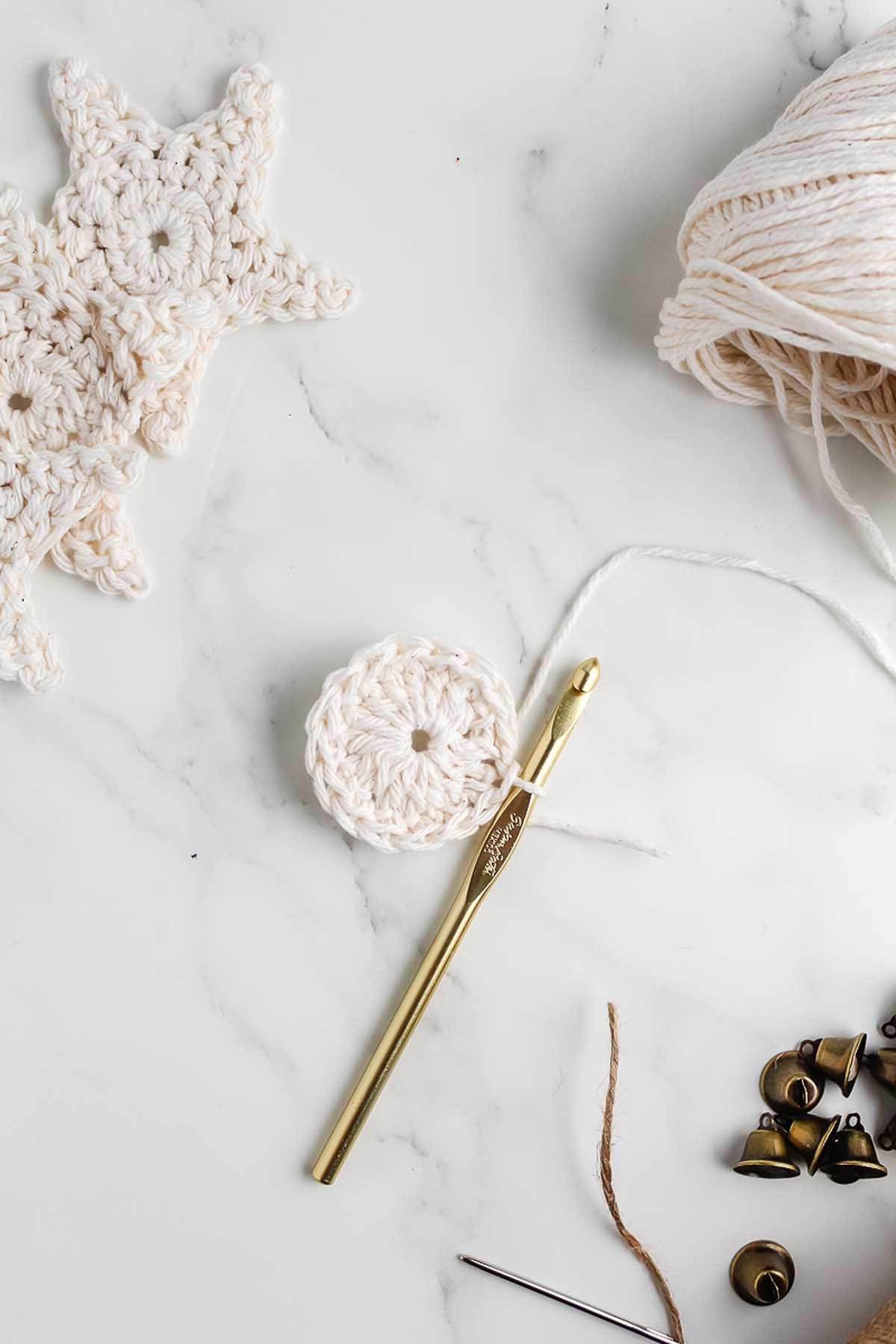 A gold crochet hook with white yarn and a round crochet piece rests on a marble surface, surrounded by star motifs—perfect inspiration for learning how to make a crochet star garland.