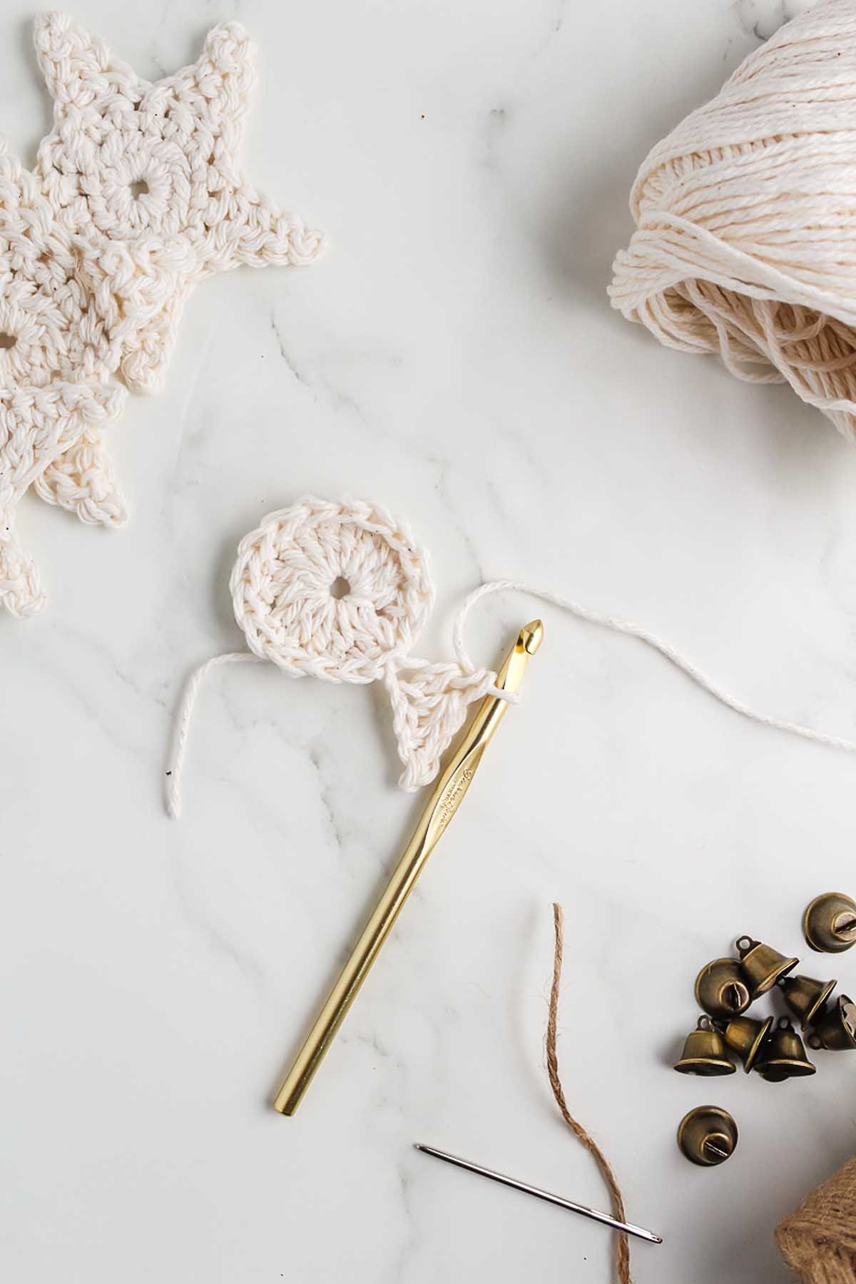 Gold crochet hook with white yarn, crocheted star, small bells, and needle on a white surface—perfect materials for learning How to Make a Crochet Star Garland.