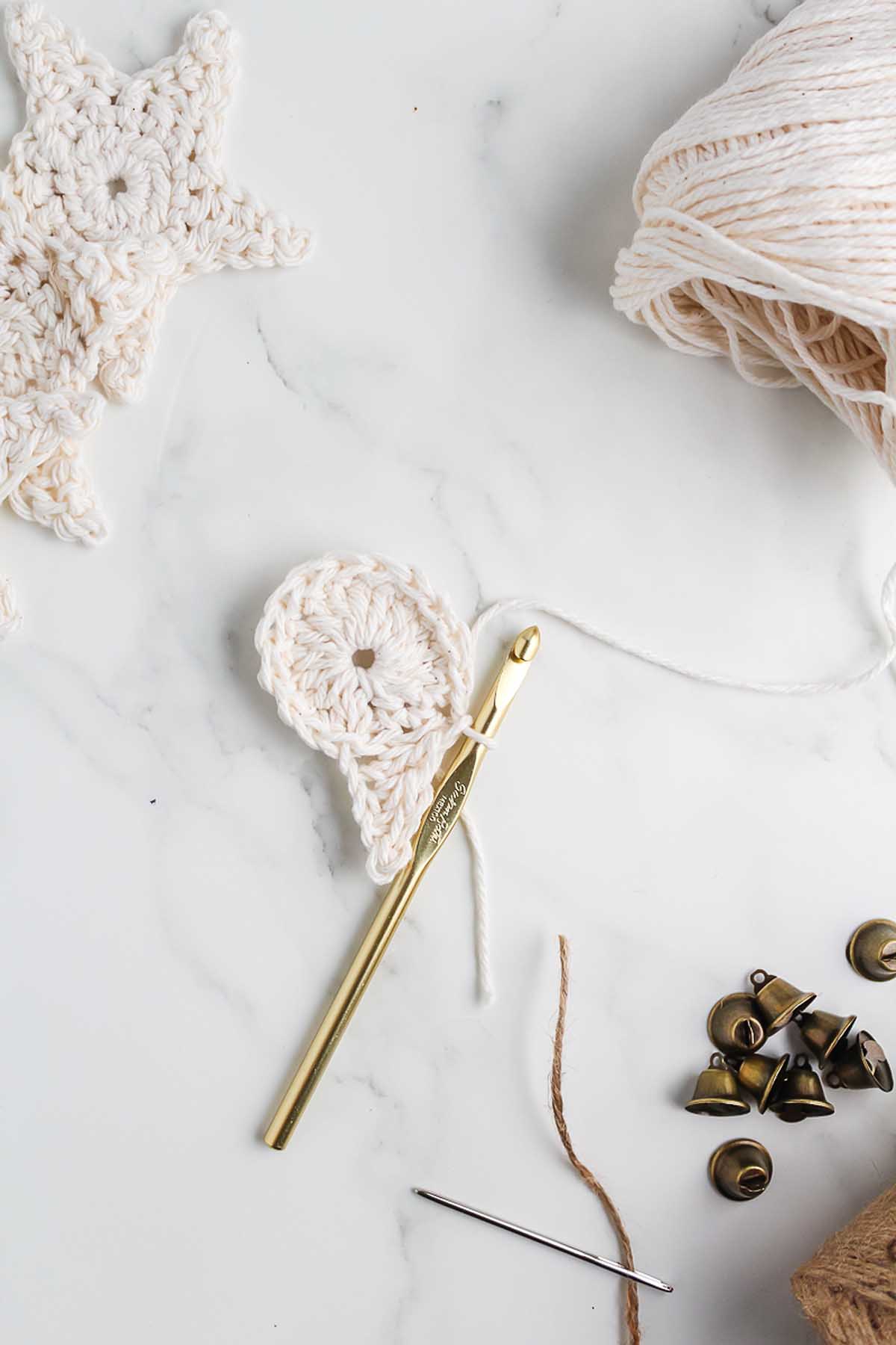 Gold crochet hook with cream yarn, star crochet piece, mini bells, and twine on a white marble surface—perfect supplies for How to Make a Crochet Star Garland.