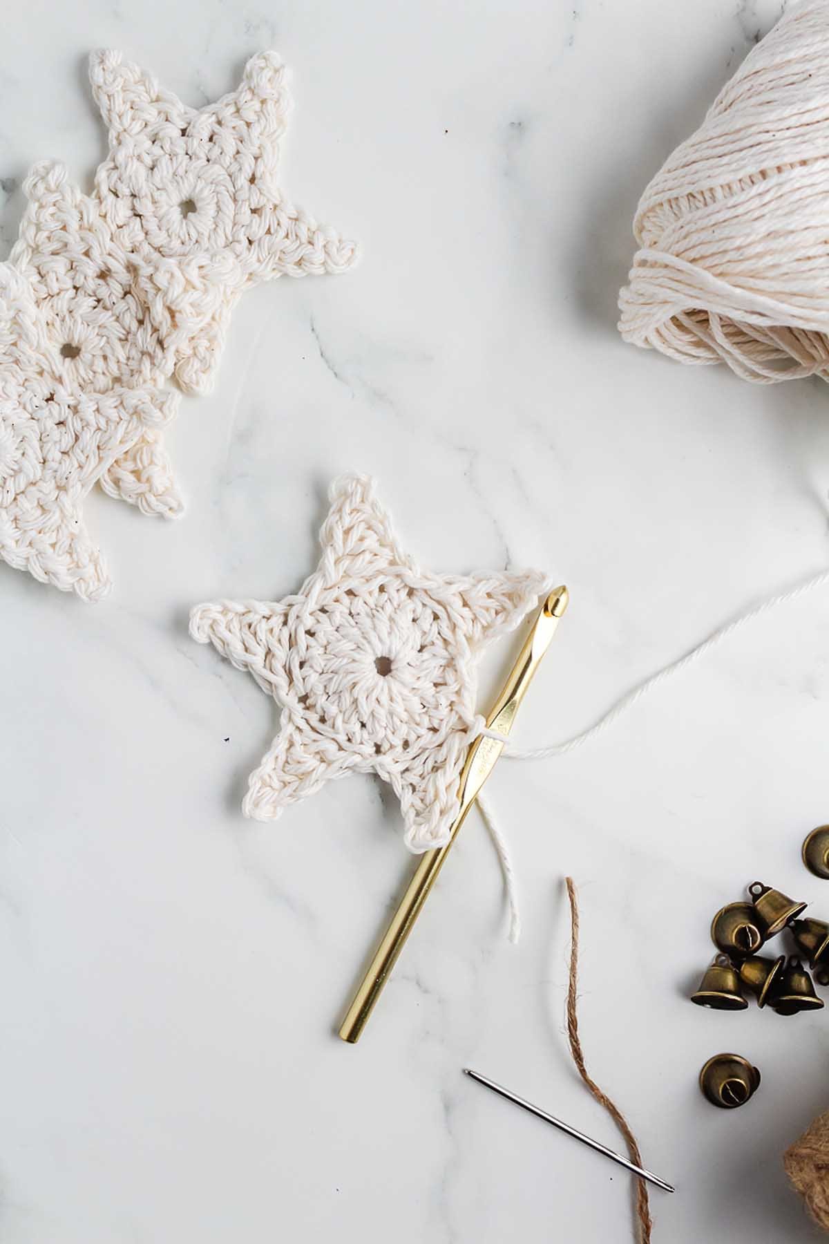 Crocheted white star with a gold hook, yarn ball, jingle bells, and needle on a marble surface—perfect supplies for How to Make a Crochet Star Garland.