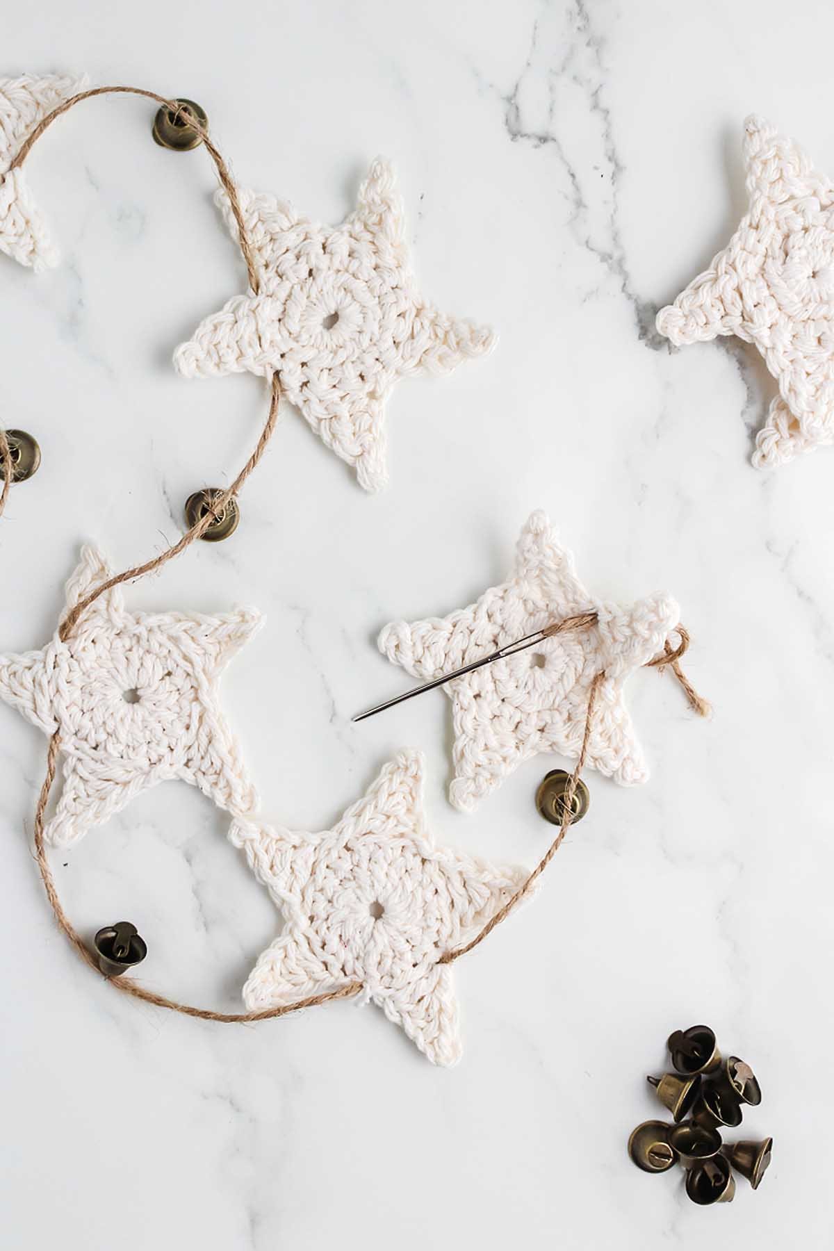 Crocheted white star garland with jingle bells and a needle on a marble surface—perfect inspiration for those learning how to make a crochet star garland.