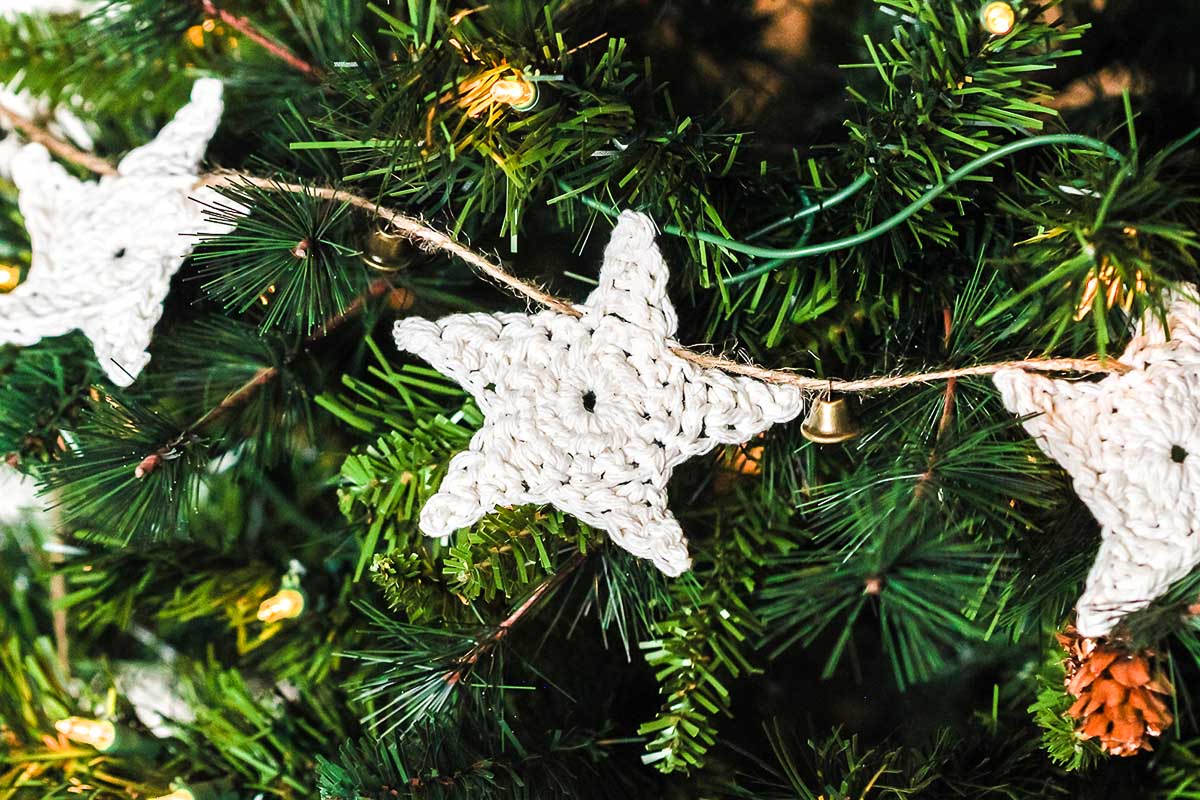 Crocheted white star ornaments, inspired by "How to Make a Crochet Star Garland," hang elegantly on a green Christmas tree with twinkling lights and a small bell.