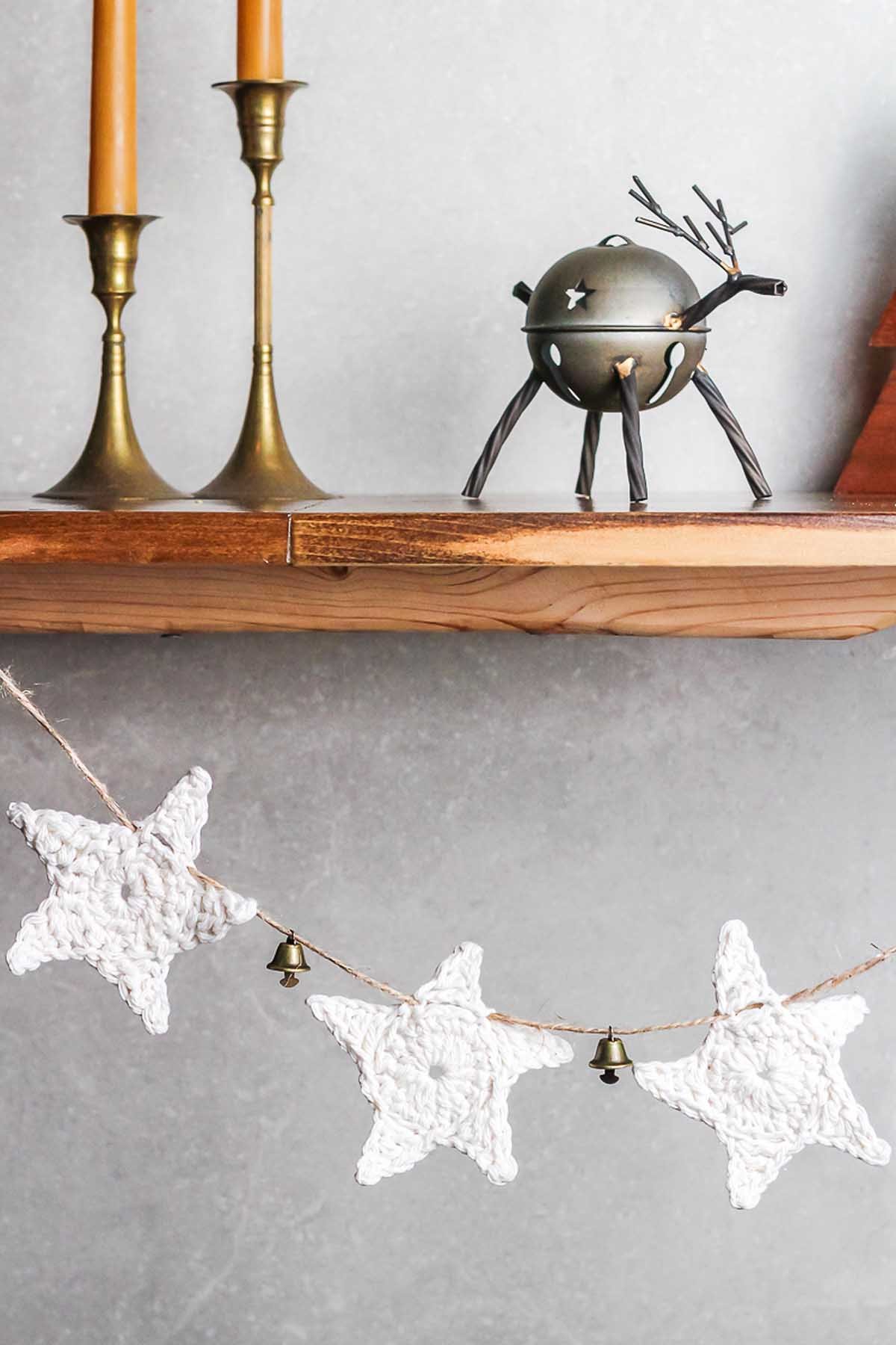 White crocheted star garland, reminiscent of DIY projects like How to Make a Crochet Star Garland, with small bells hangs below a wooden shelf adorned with candles and a reindeer figurine.