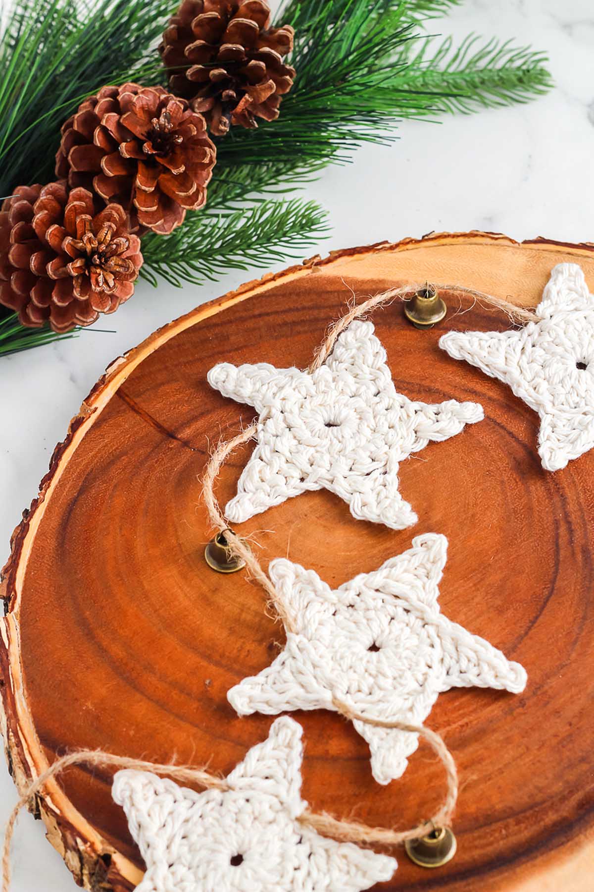 Crocheted white star garland on a wooden tray, with pinecones and pine branches in the background—perfect for those searching how to make a crochet star garland for festive decor.