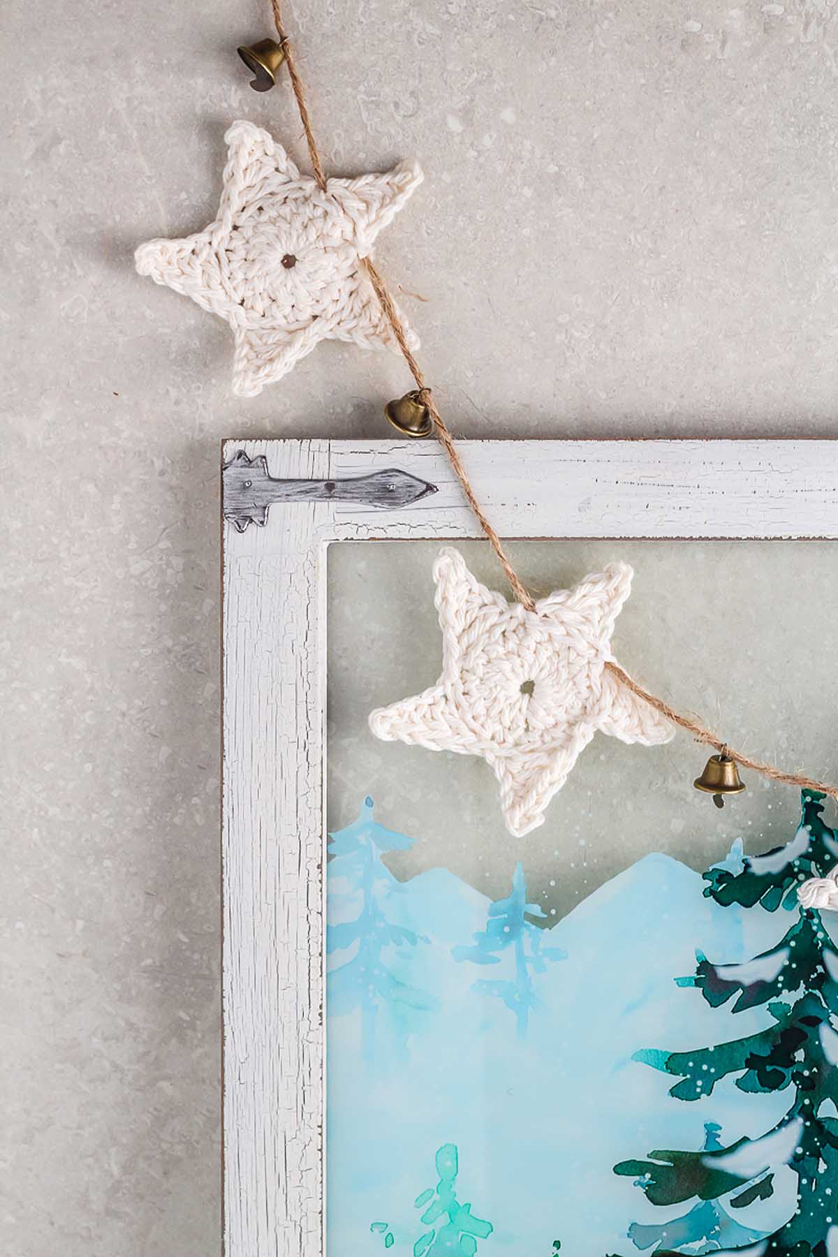Crocheted star garland with small bells, inspired by How to Make a Crochet Star Garland, hanging on a corner of a rustic white frame with winter scene art.