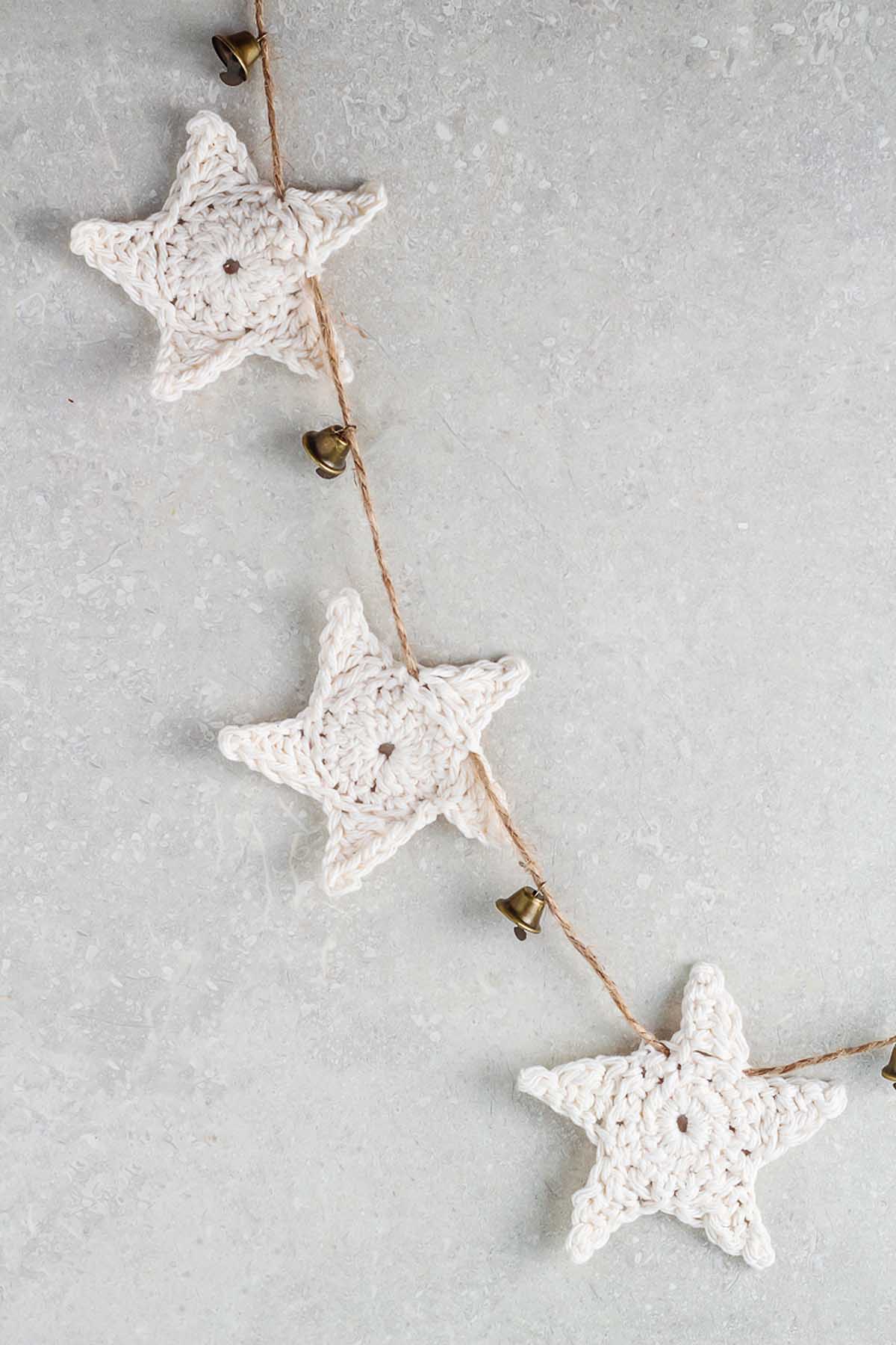 String of crocheted white star ornaments with small bells on a light gray background, perfect for anyone learning how to make a crochet star garland.