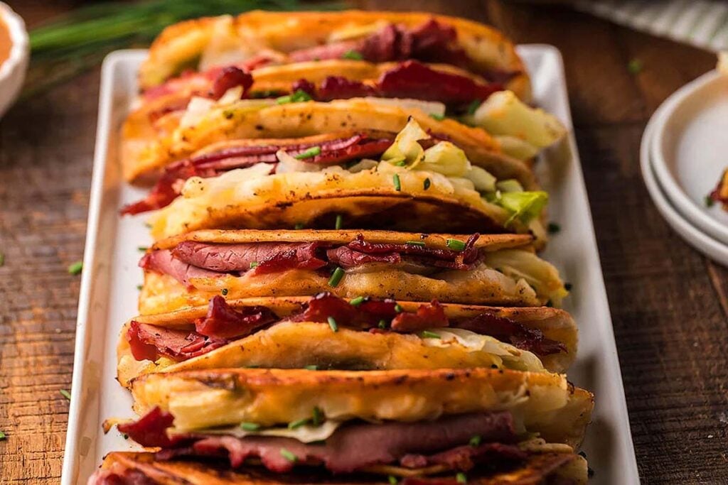 A row of crispy tacos filled with corned beef, cabbage, and chives on a rectangular white plate—perfect for those seeking unique taco recipes.