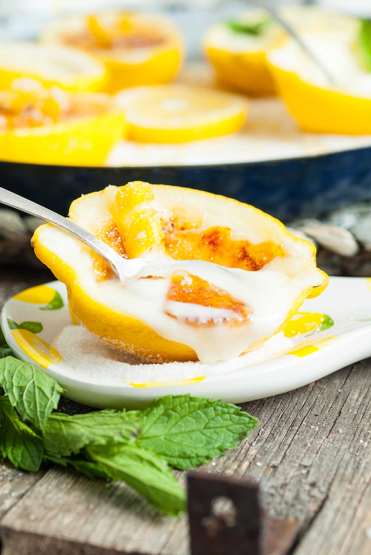 A Lemon Posset Brûlée: a lemon half filled with caramelized sugar and cream, served with a spoon on a plate garnished with mint leaves.