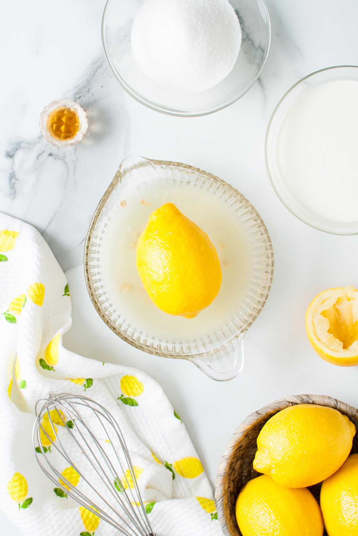 Lemon Posset Brûlée ingredients: lemon on a juicer, whole lemons, sugar, milk, a whisk, and a lemon towel arranged on a white surface.