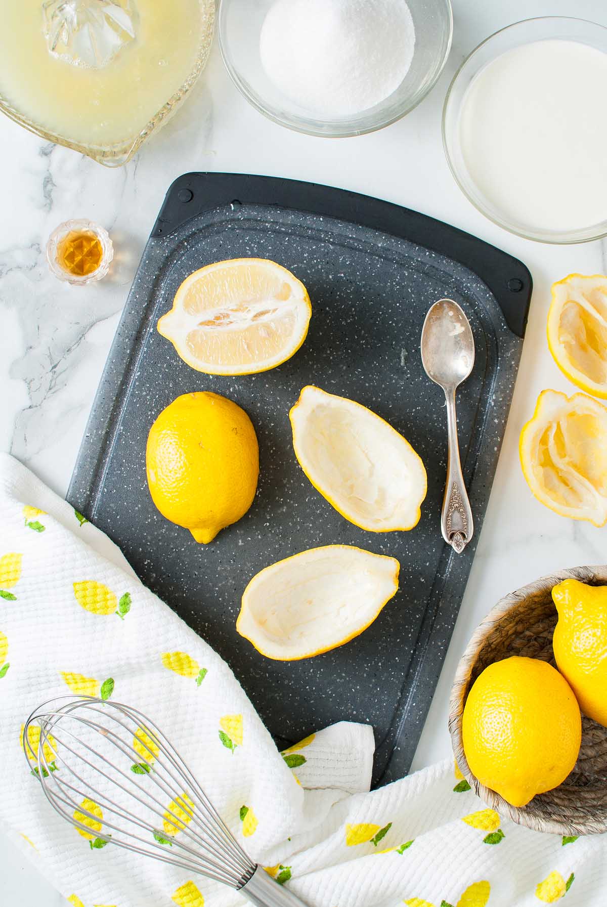 Lemons, halved and hollowed for a delicious Lemon Posset Brûlée, rest on a cutting board with a spoon, surrounded by ingredients and utensils.