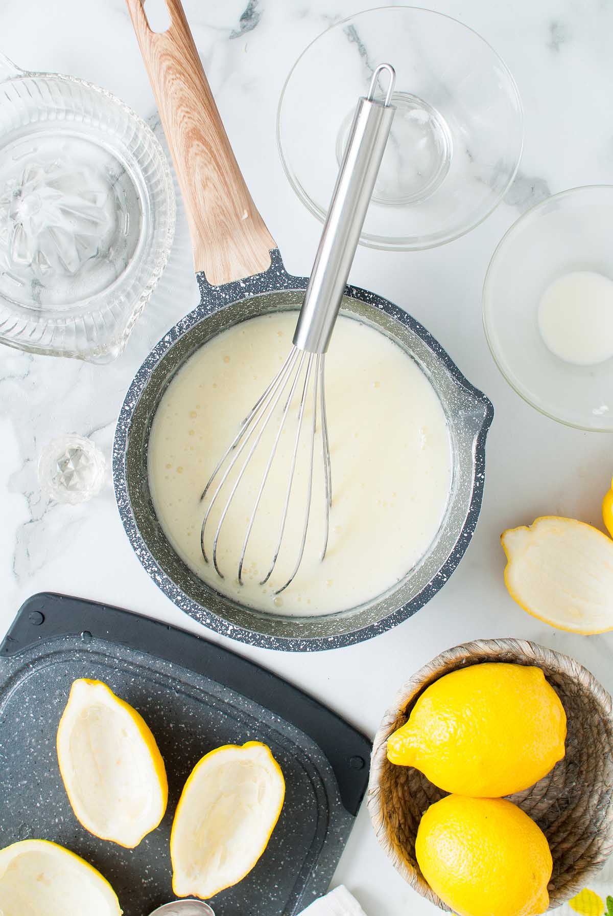 A whisk in a saucepan of creamy mixture, surrounded by lemons, lemon halves, and bowls on a marble surface—perfect for preparing a luscious Lemon Posset Brûlée.
