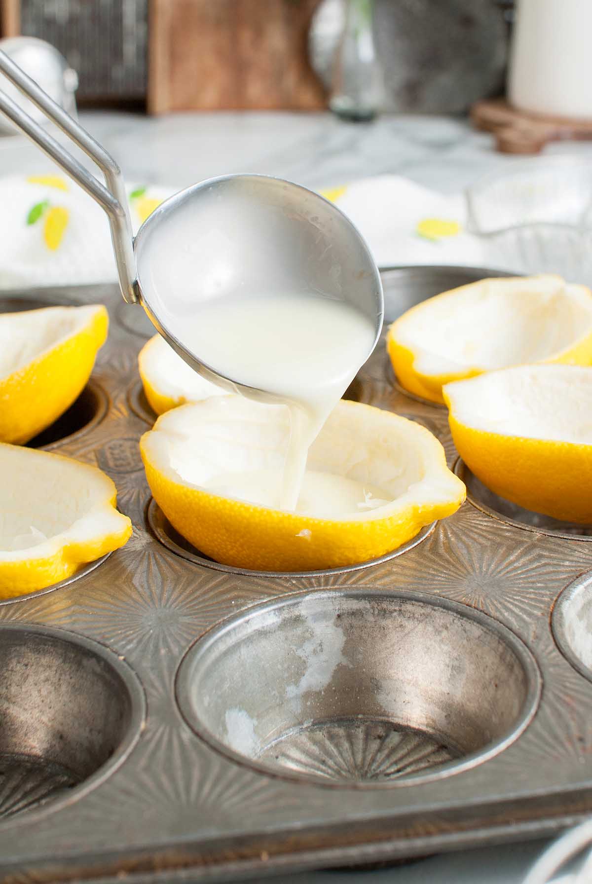 A metal scoop pours white liquid into a hollowed lemon half set in a muffin tin, preparing it for Lemon Posset Brûlée.