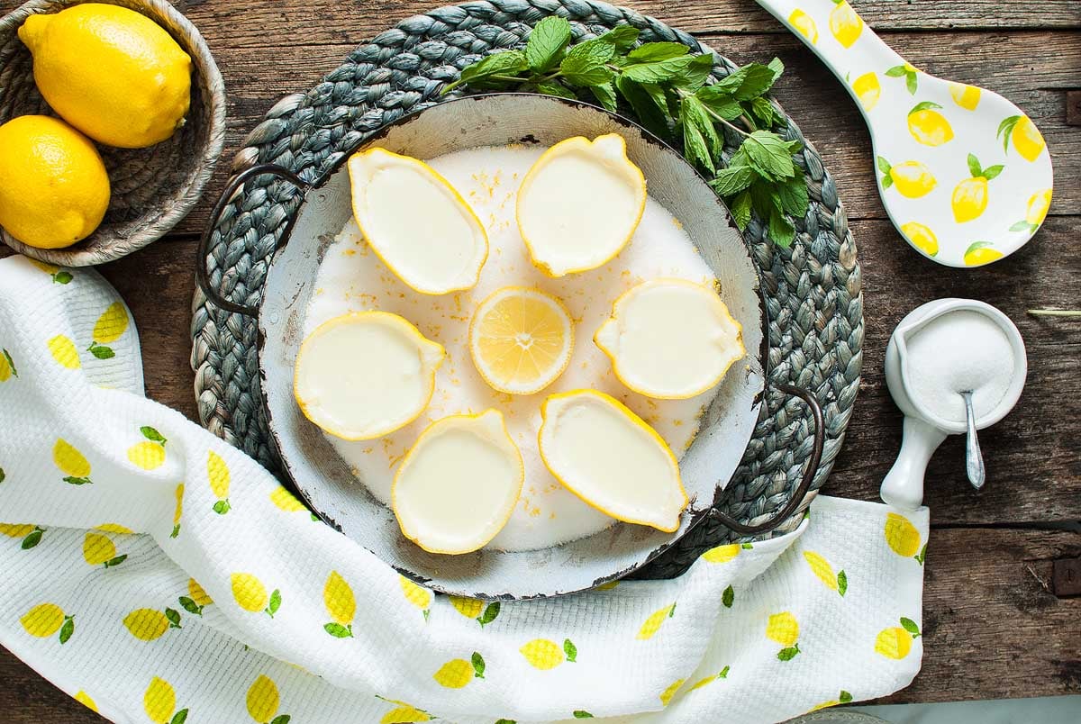 Six Lemon Posset Brûlée desserts shaped like lemons sit on a tray, surrounded by fresh lemons, mint, and lemon-themed kitchen accessories.