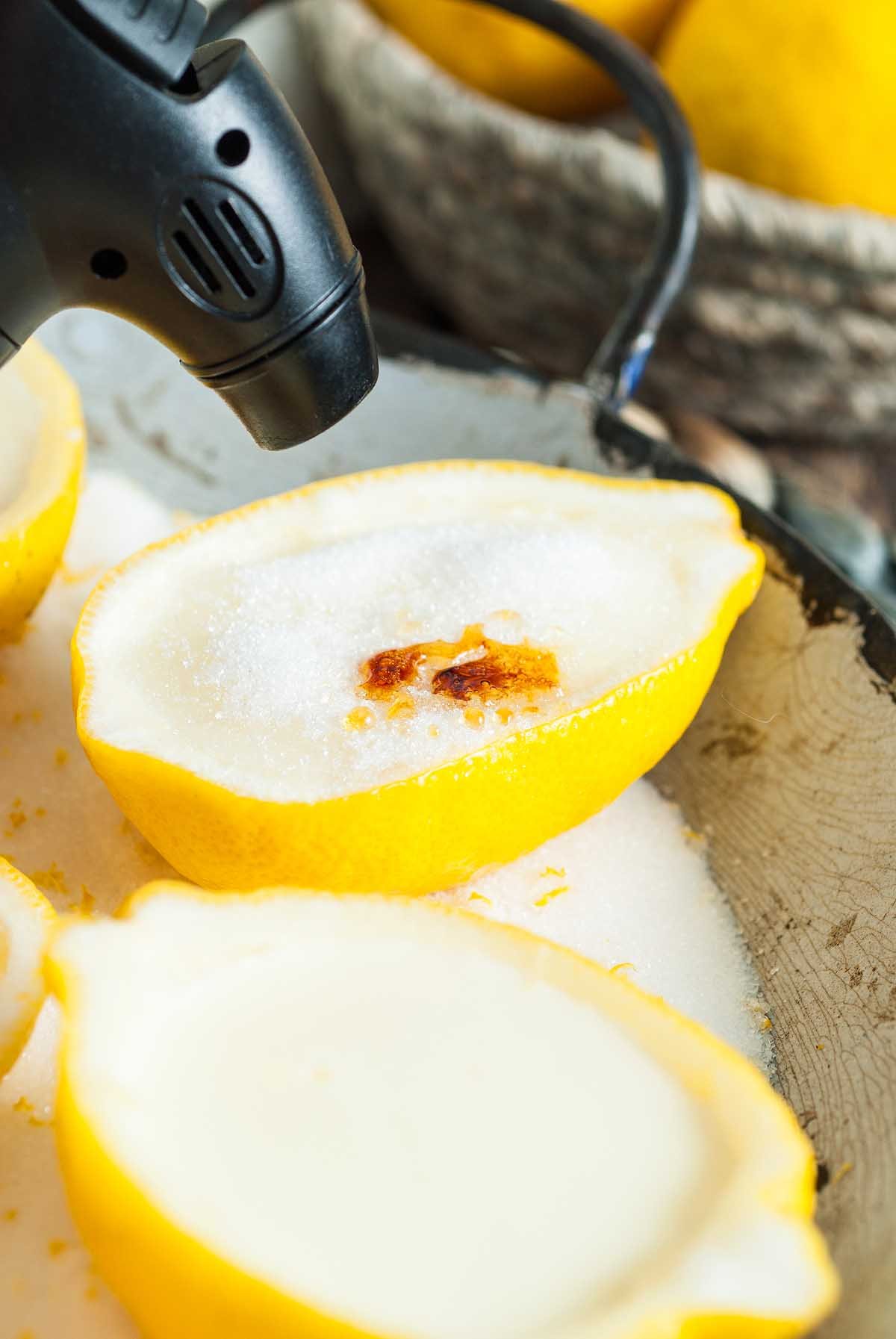 A kitchen torch caramelizes sugar on a lemon half filled with sugar, creating a Lemon Posset Brûlée-inspired treat.