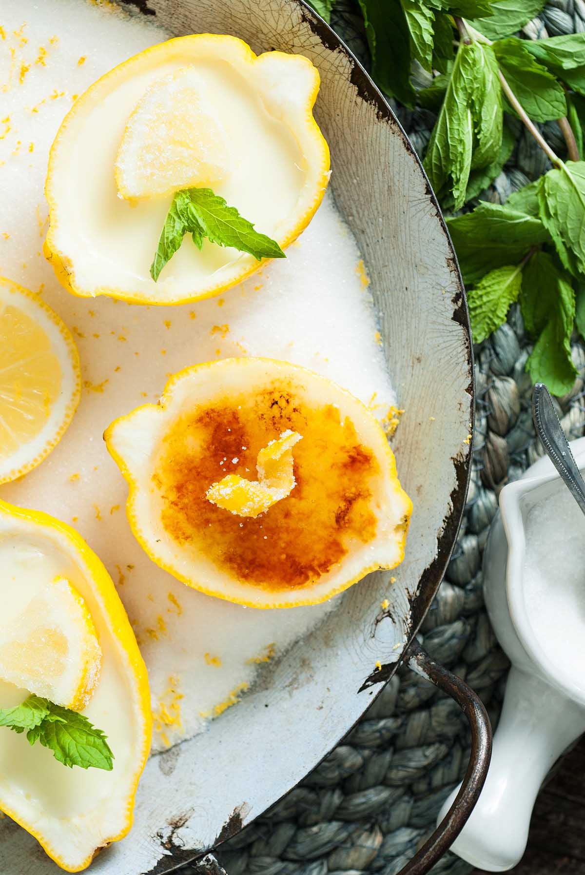 Lemon Posset Brûlée served in lemon halves, one perfectly brûléed, filled with creamy dessert and garnished with mint and sugar on a rustic tray.