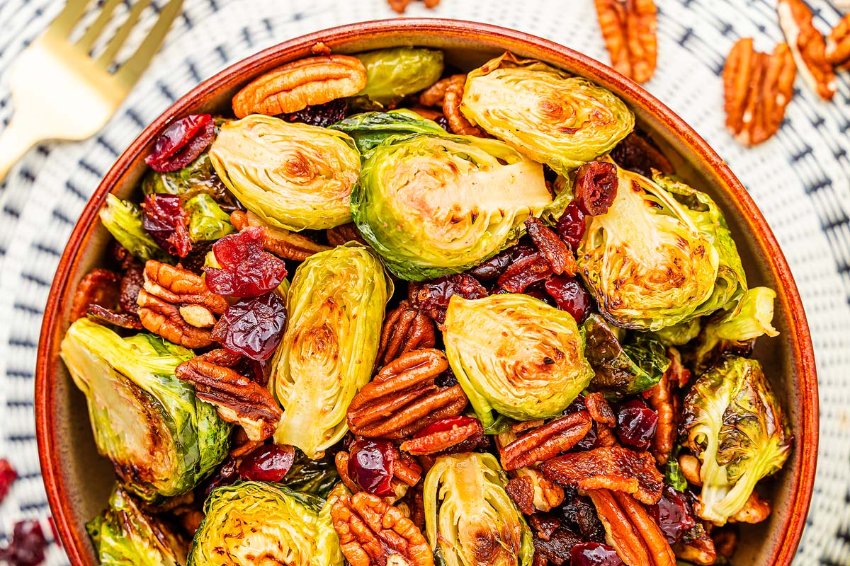 A close-up of a bowl filled with roasted Brussels sprouts, pecans, and dried cranberries. A fork is partially visible in the background, resting on a woven, checkered placemat. The vibrant colors of the dish stand out, highlighting the golden brown and green textures.