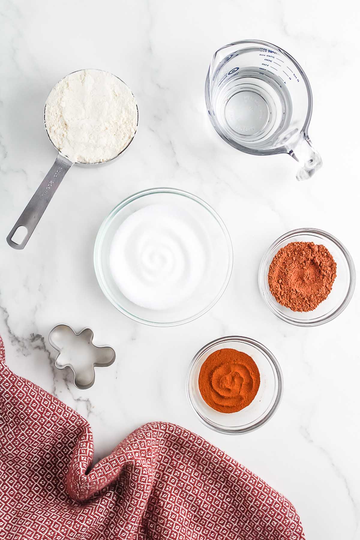 Baking ingredients in bowls and a measuring cup on a white surface, with a cookie cutter and red towel&mdash;perfect for creating a Salt Dough Gingerbread Man Garland.