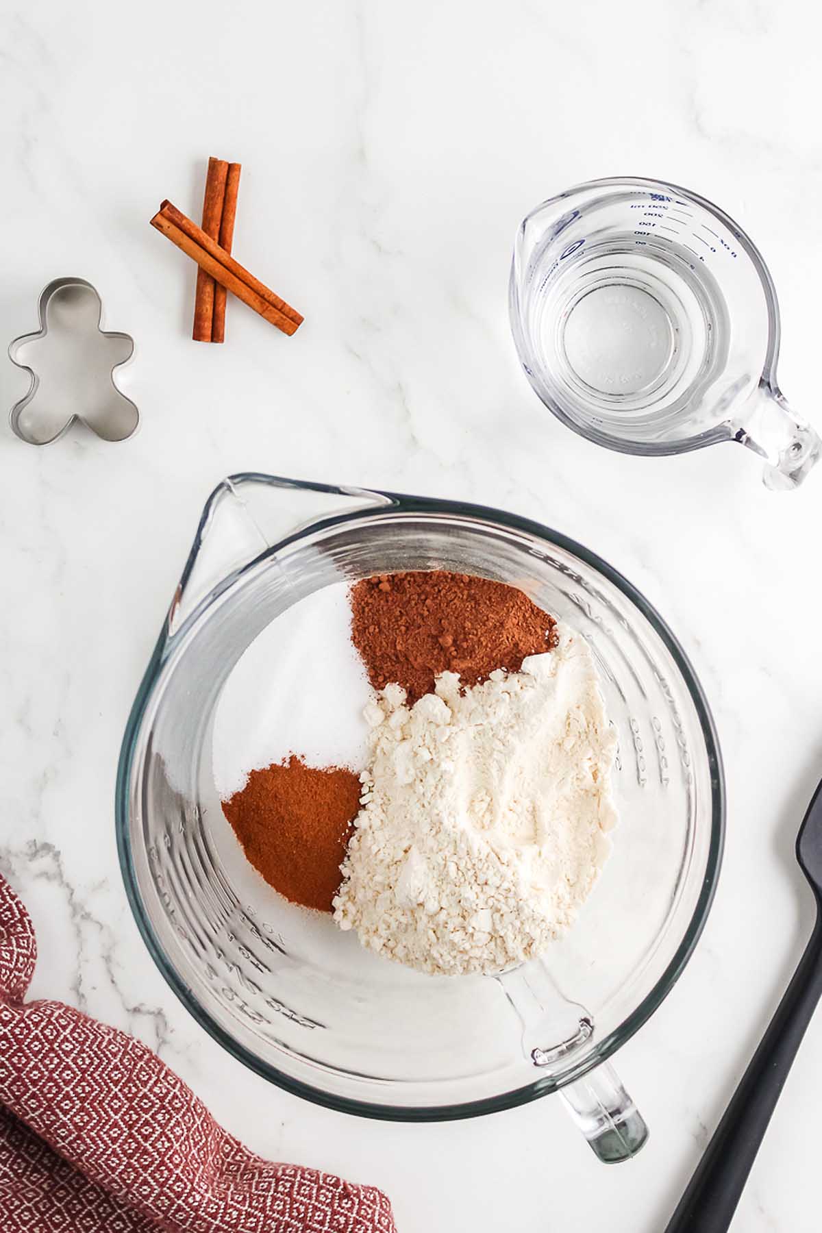 Glass bowl with flour, sugar, and spices sits beside a measuring cup of water, cinnamon sticks, a gingerbread cutter, and ingredients for a Salt Dough Gingerbread Man Garland.