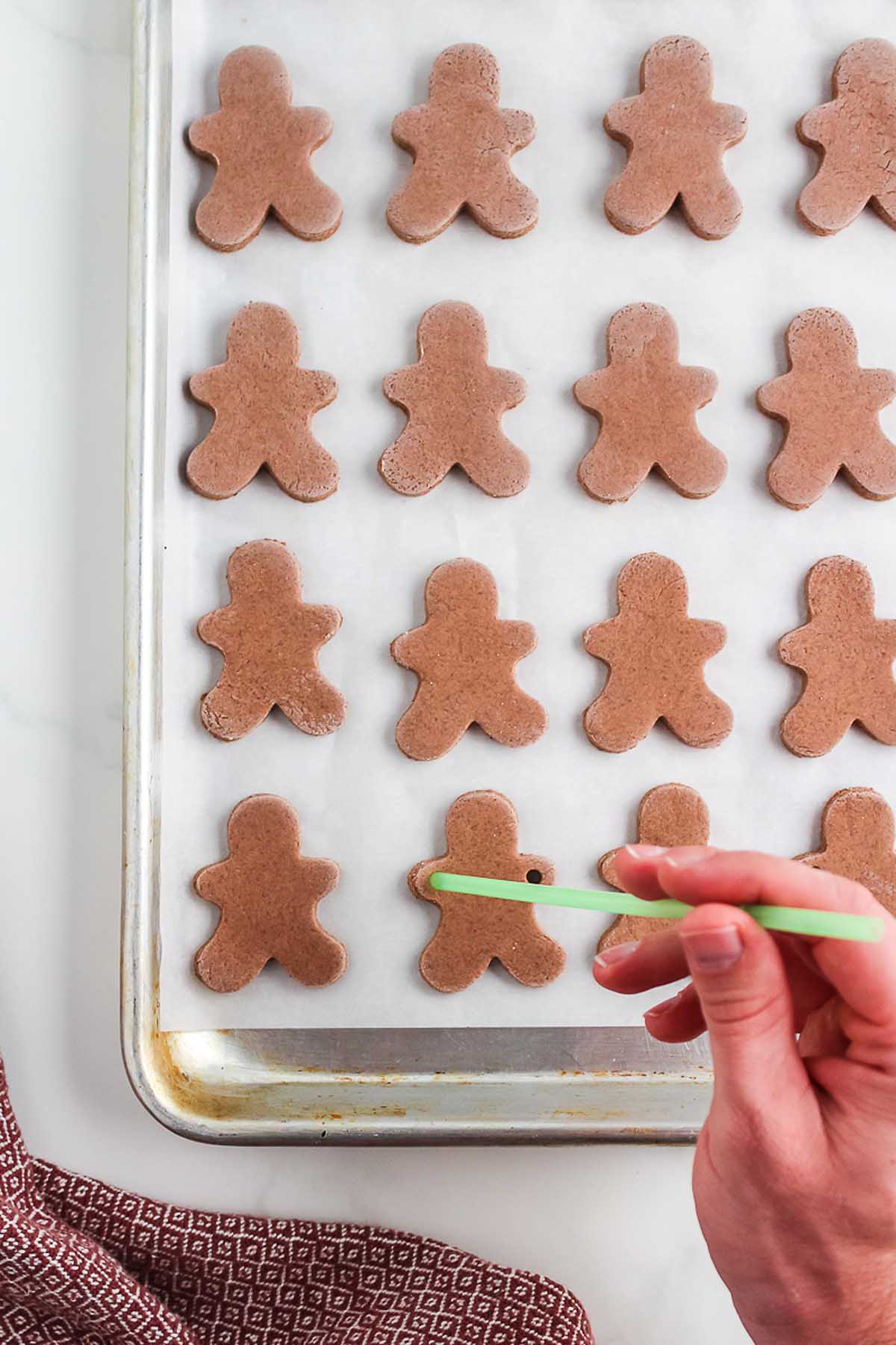 A hand uses a straw to make holes in chocolate gingerbread men cookies on a baking tray, perfect for creating a Salt Dough Gingerbread Man Garland.
