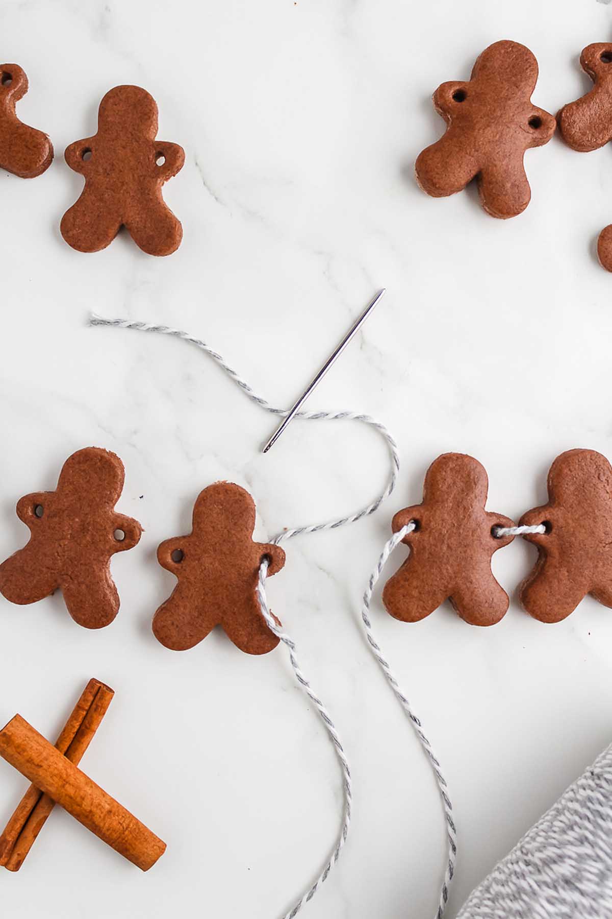 Salt Dough Gingerbread Man Garland ornaments are being threaded with string and a needle, while cinnamon sticks rest nearby, adding a festive touch.