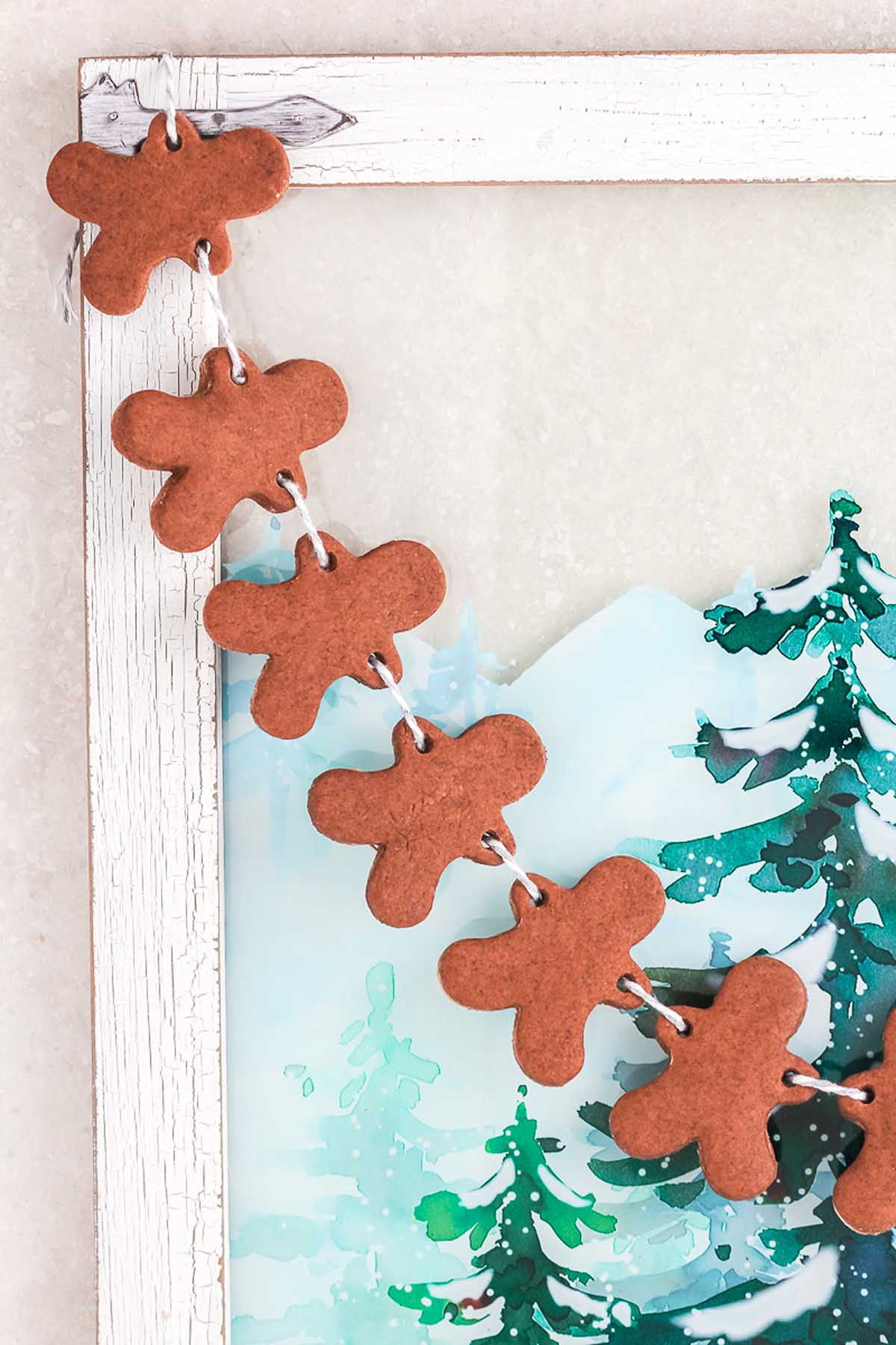 Salt Dough Gingerbread Man Garland draped along a rustic white wooden frame, featuring a charming winter tree illustration.