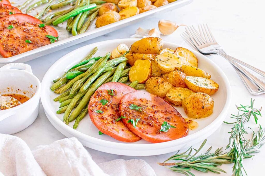 White plate with roasted potatoes, green beans, and slices of ham topped with herbs&mdash;this Sheet Pan Ham Dinner is served on a light background.