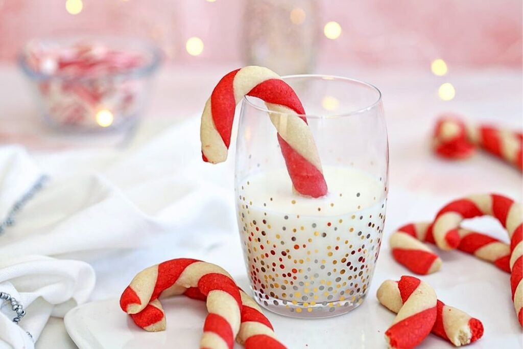A candy cane cookie dipped into a glass of milk with candy cane cookies around.