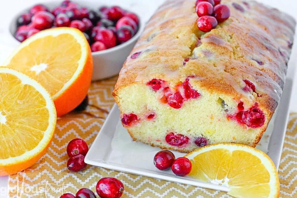 A loaf of orange cranberry bread with oranges and cranberries around.