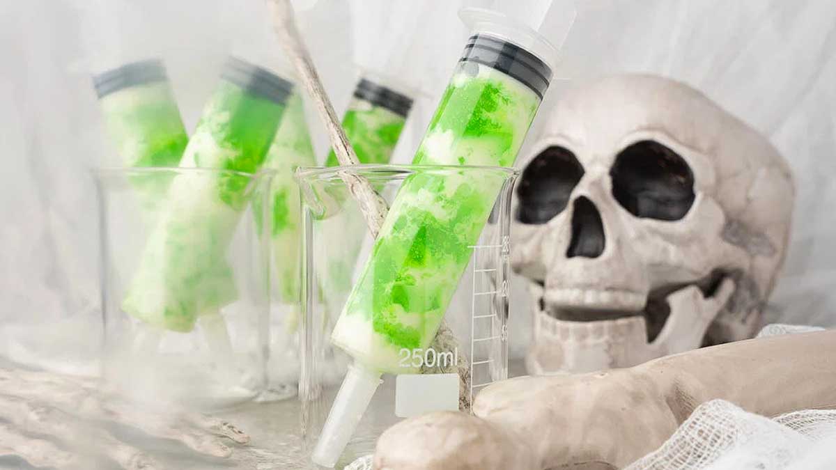 Green syringes in beakers with a skull and bones in the background, creating a spooky laboratory scene perfect for Halloween jello shots.