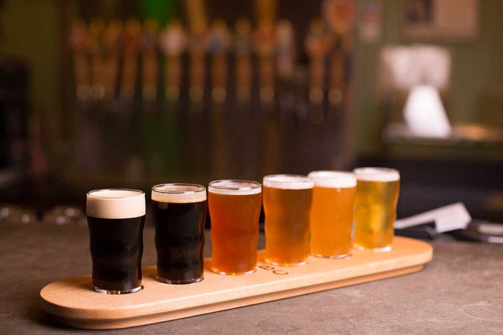 Close up of craft beer tasting flight at local brewery of small pint glasses in a row on a tray with rainbow variety of dark malt stouts to golden yellow hoppy ales on bar, with taps in background.