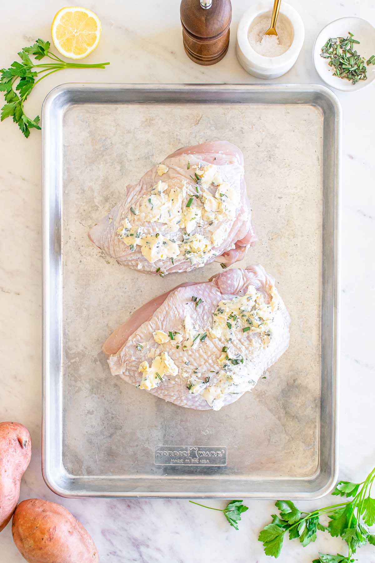 Two turkey breasts on a baking sheet with potatoes, herbs, and stuffing.