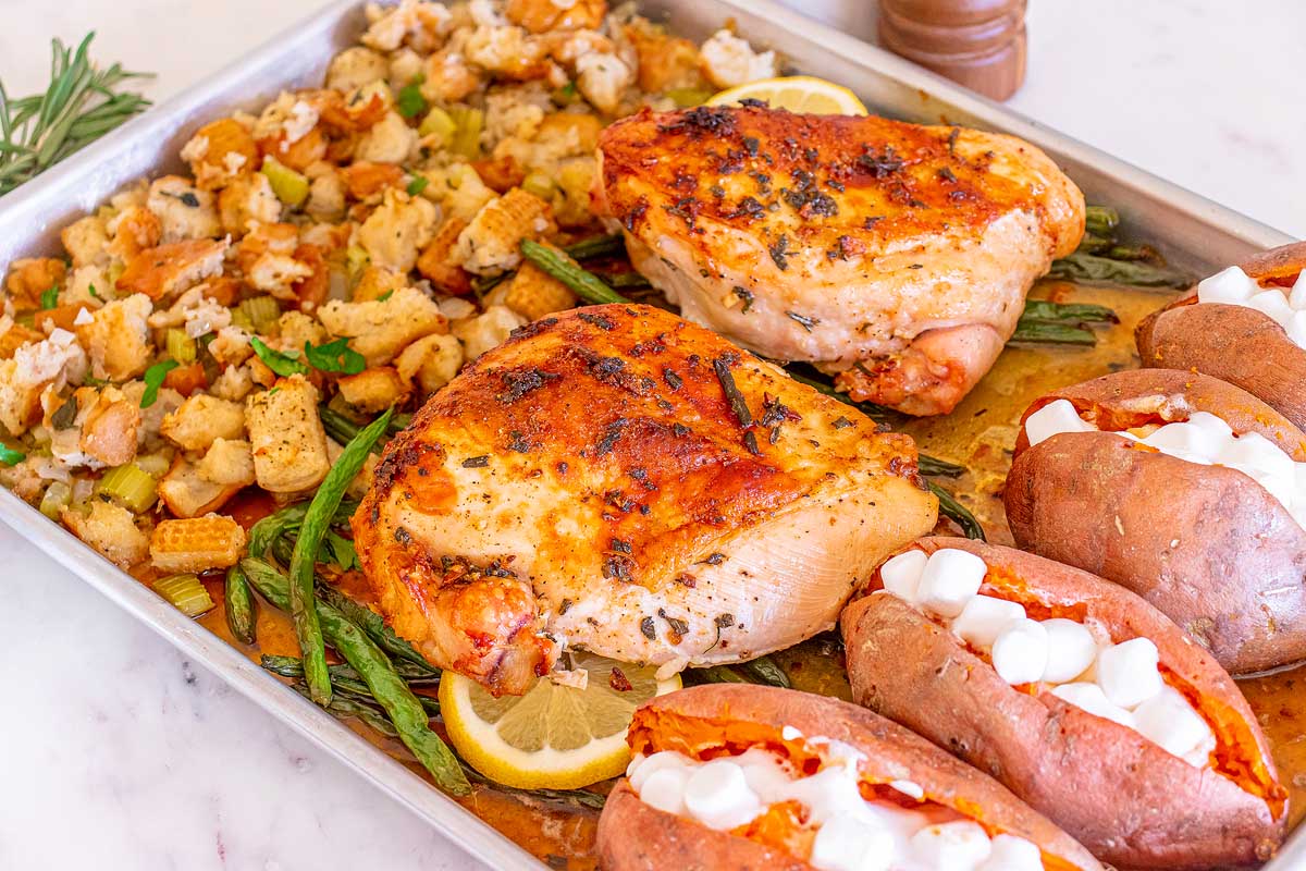A sheet pan meal with roasted chicken breasts, stuffing, green beans, and twice-baked sweet potatoes topped with melted marshmallows. Lemon slices and herbs garnish the dish. All ingredients are arranged on a baking sheet on a light countertop.