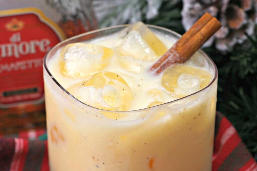 A creamy drink inspired by classic eggnog recipes, served over ice cubes with a cinnamon stick garnish in a glass set atop a festive cloth.