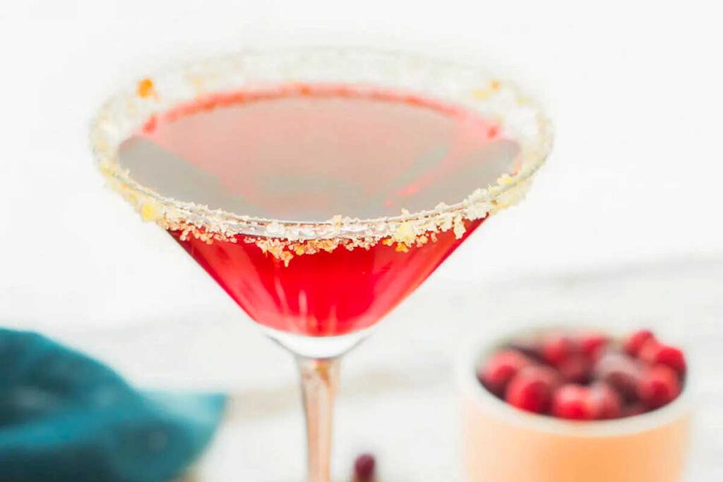 A red cocktail in a martini glass with a sugared rim; perfect for Friendsgiving, with a bowl of cranberries in the background.
