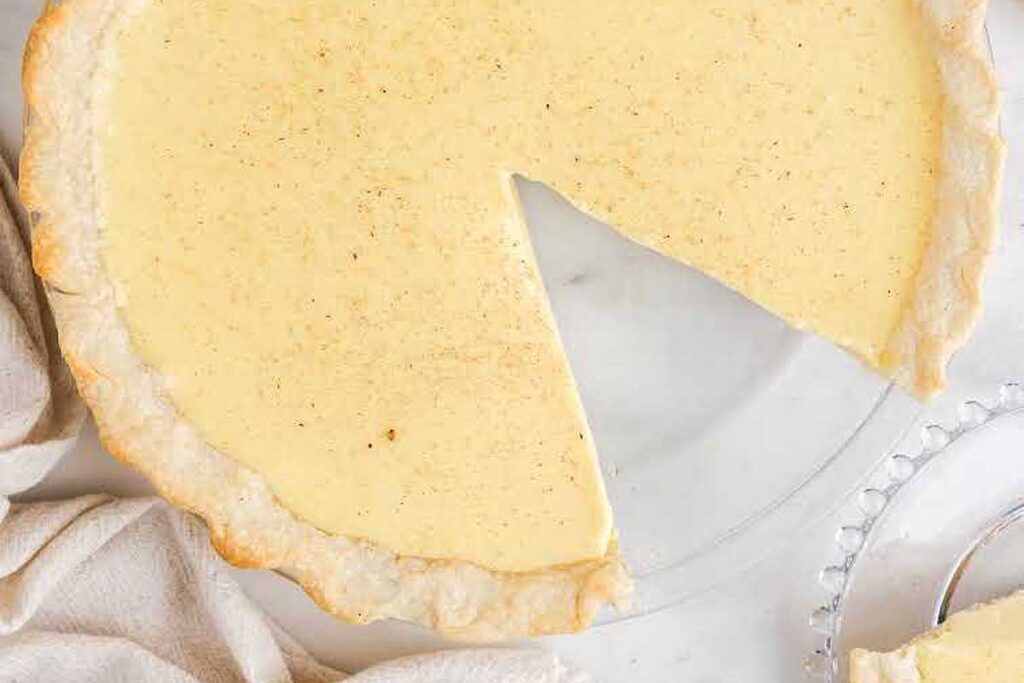 A creamy custard pie in a flaky crust with one slice removed, shown from above on a white surface—perfect for fans of classic eggnog recipes.