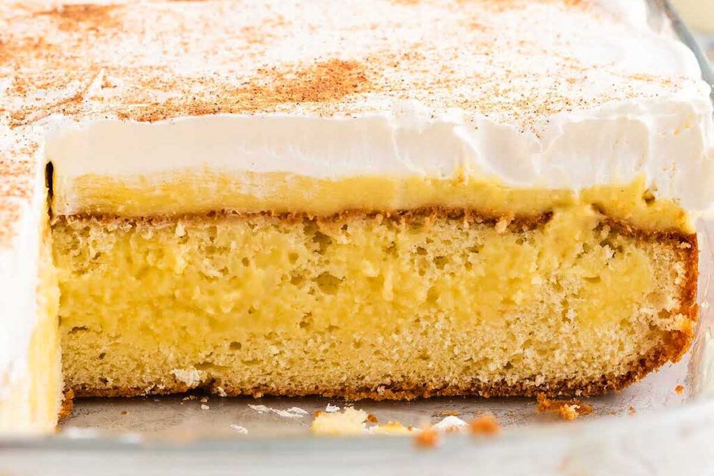 Close-up of a sliced cake with creamy yellow filling and a thick layer of whipped topping, dusted with cinnamon—perfect for fans of festive eggnog recipes.