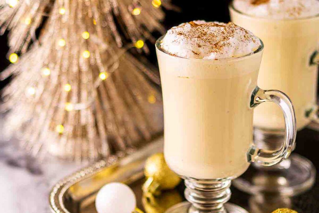 A glass mug of eggnog, inspired by classic eggnog recipes, topped with whipped cream and cinnamon, sits next to a festive tree and ornaments.