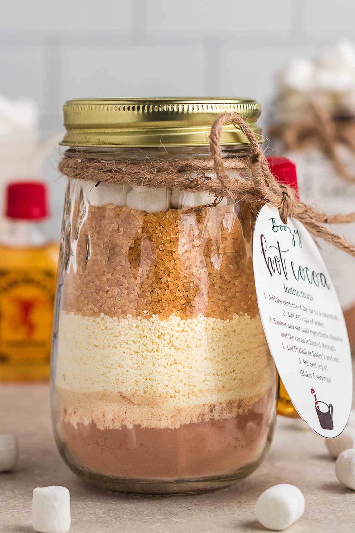 A mason jar filled with layered hot cocoa mix, topped with marshmallows, and tied with a recipe tag—this Boozy Hot Chocolate Gift Kit makes a delightful treat for cozy nights in.