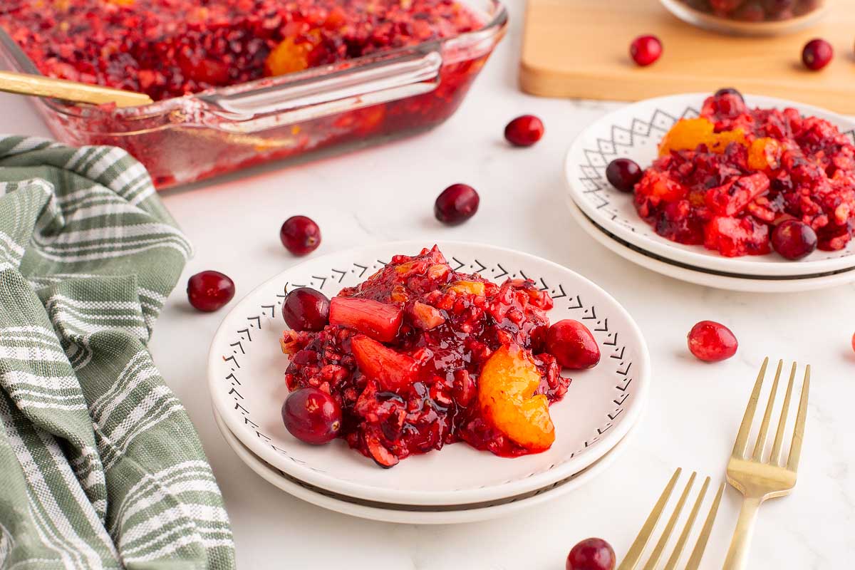 A white plate features a serving of vibrant cranberry salad with whole cranberries and orange slices, set on a white table next to gold forks, a green and white cloth, and another plate of salad in the background. A glass baking dish with more salad is also visible.