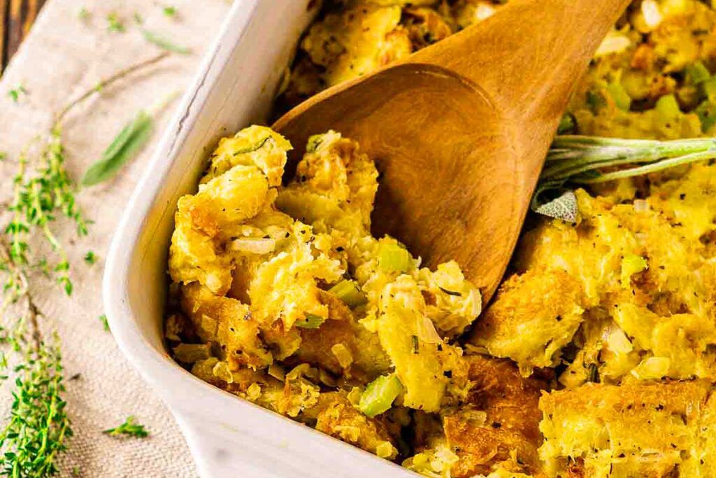 A wooden spoon scooping homemade stuffing with celery from a white baking dish—perfect for serving at your next Friendsgiving gathering.