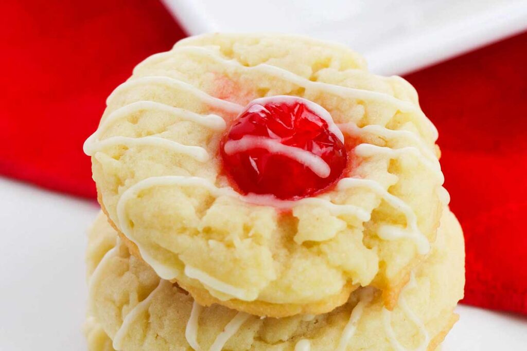A stacked cookie with a red cherry center and white icing drizzle, on a red and white background—perfect for pairing with your favorite eggnog recipes.