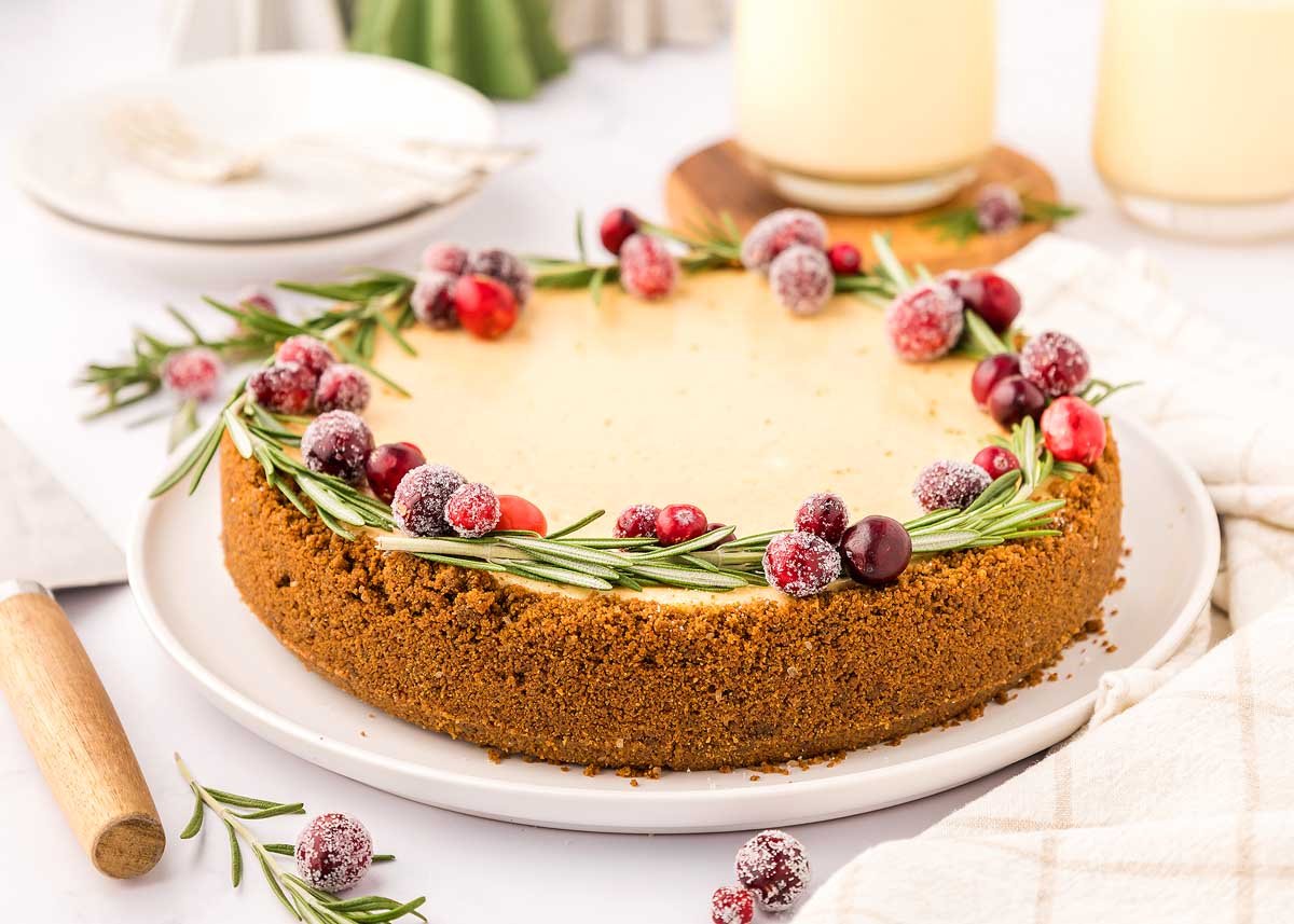 A festive cheesecake with cranberries and sprigs of rosemary on a white plate.
