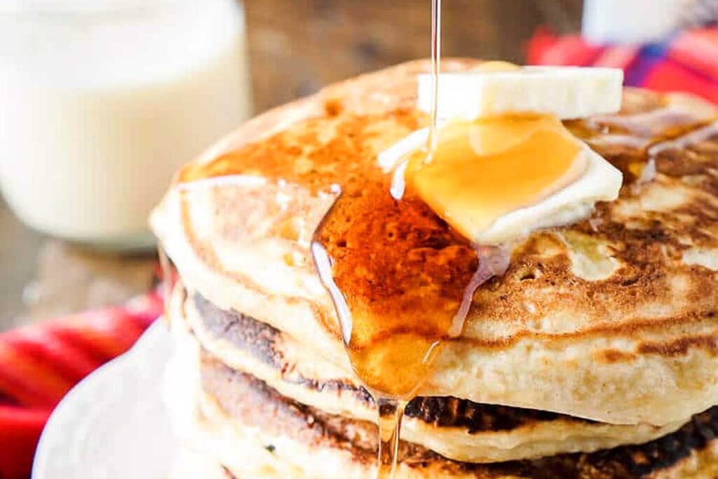 Stack of pancakes topped with butter and syrup being poured, with a glass of milk in the background—perfect for pairing with classic eggnog recipes.