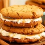 Two Gingerbread Sandwich Cookies filled with creamy frosting and caramel, stacked on a wooden board with cinnamon sticks.