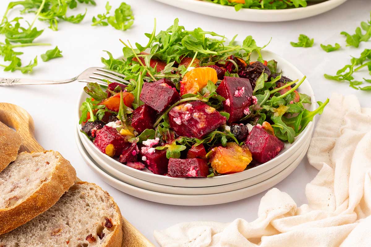 A colorful salad with roasted red beets, orange segments, arugula, and feta cheese, served in a white bowl. A fork rests on the edge of the bowl. Pieces of rustic bread sit on a wooden board next to the bowl. Some arugula leaves are scattered around the plate.