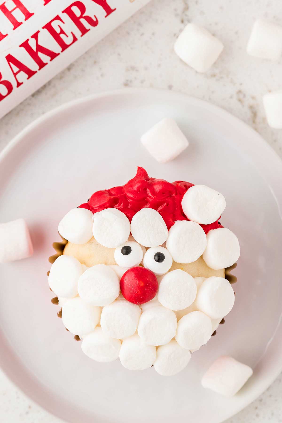 A festive Christmas cupcake adorned with a jolly Santa face, complete with marshmallow features, presented on a plate.